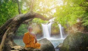 A monk sitting on the rocks near a waterfall.